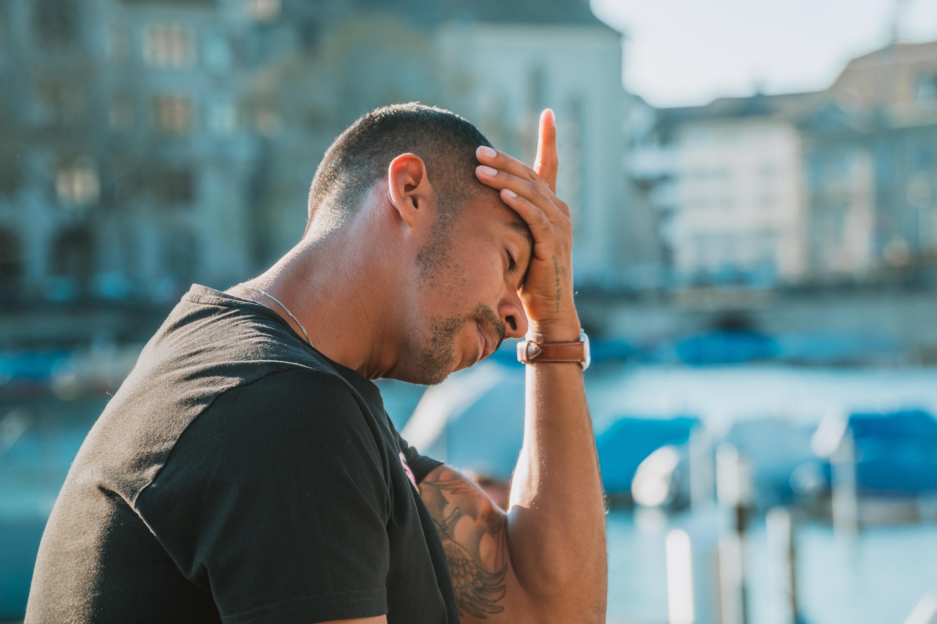 Vakantiestress: als ontspanning juist spanning wordt Een persoon staat buiten met de ogen gesloten en één hand tegen het voorhoofd, alsof hij zich overweldigd of gestrest voelt. Op de achtergrond zijn een stad en water vaag zichtbaar.